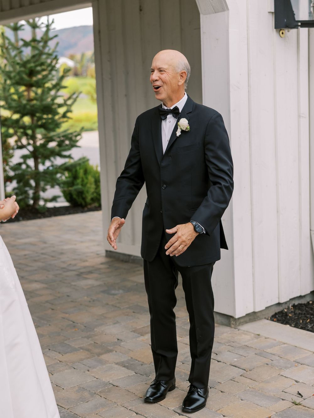 Smiling man in black tuxedo with white boutonniere stands on stone patio outside white building at wedding.