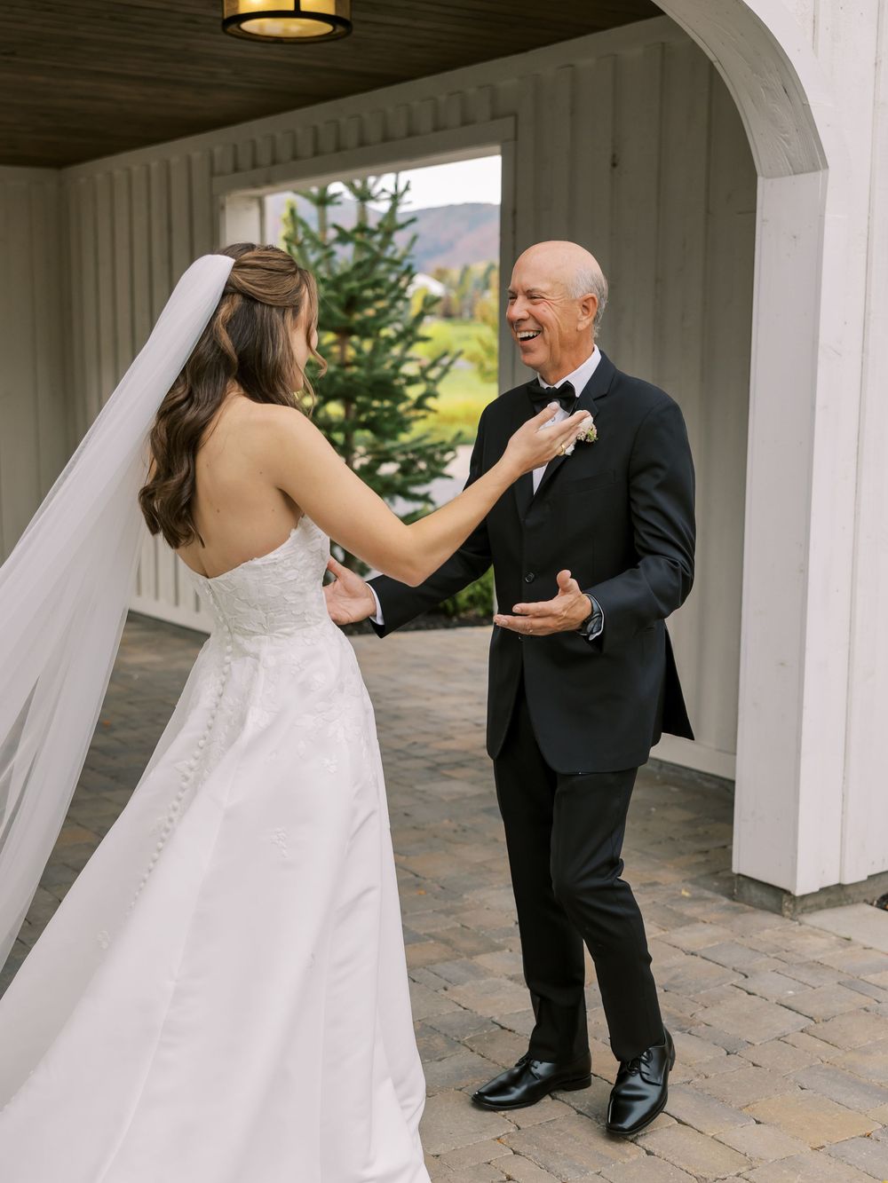 Bride in white strapless gown shares joyful first look moment with father in tuxedo at elegant wedding venue.