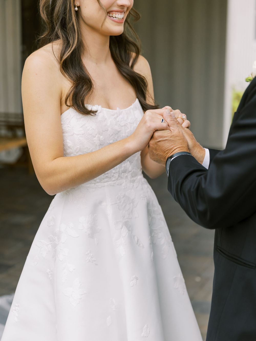 Bride in strapless white lace gown smiles while holding hands with a guest in a dark suit at her wedding.