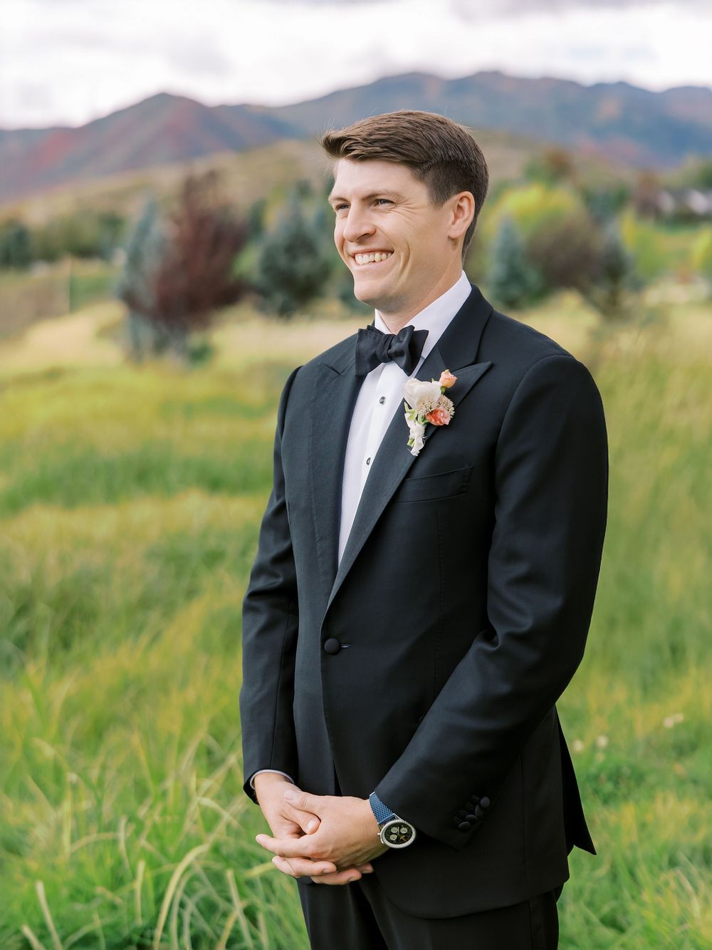 Smiling groom in black tuxedo with floral boutonniere stands outdoors with mountain landscape backdrop.