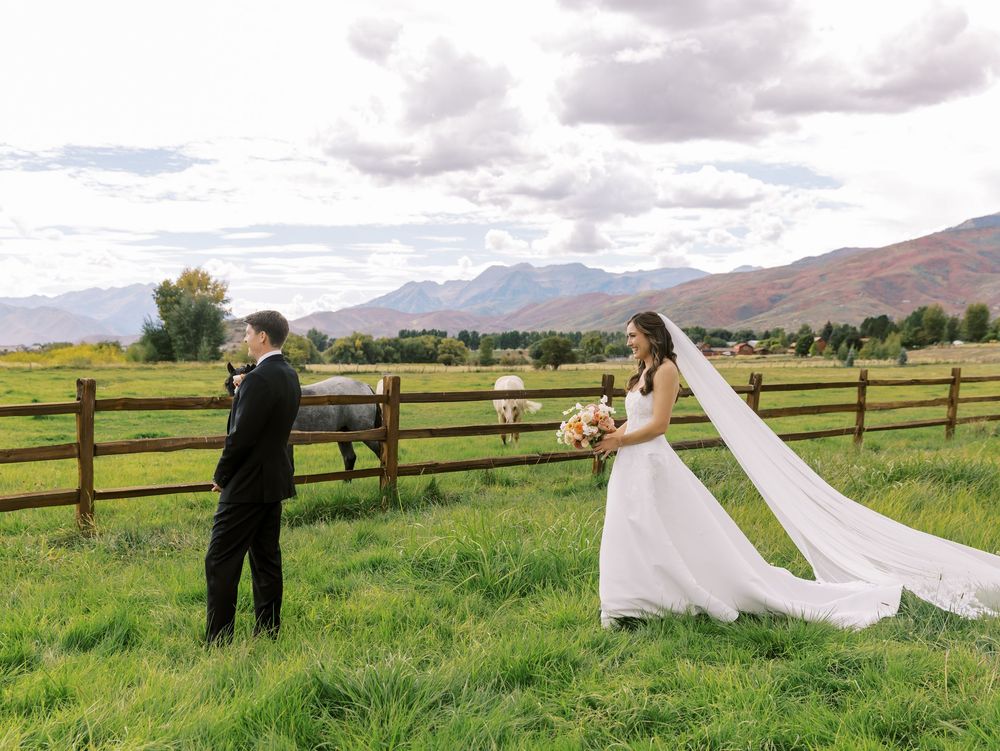 Bride and groom stand by rustic wooden fence in green meadow with mountain backdrop on their wedding day.