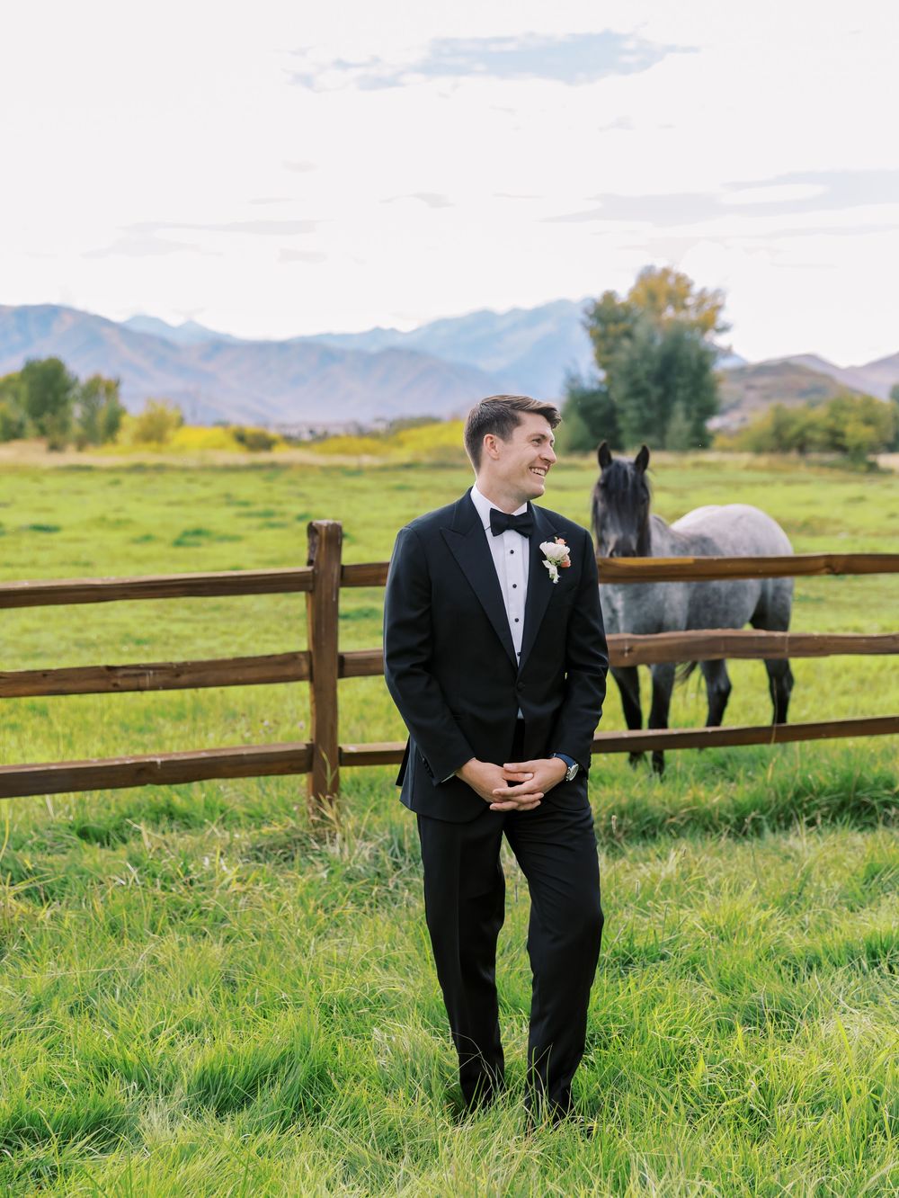 Groom in black tuxedo smiling in green pasture with horse and mountain backdrop.