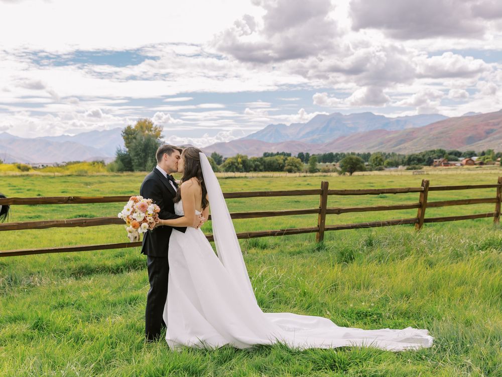 Bride and groom kissing in a green meadow with mountain views and a wooden fence on their wedding day.