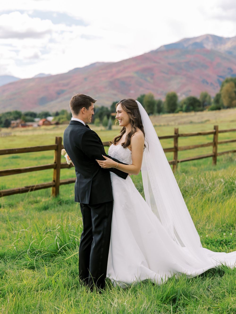 Bride and groom embrace in green field with stunning red mountain backdrop and wooden fence.