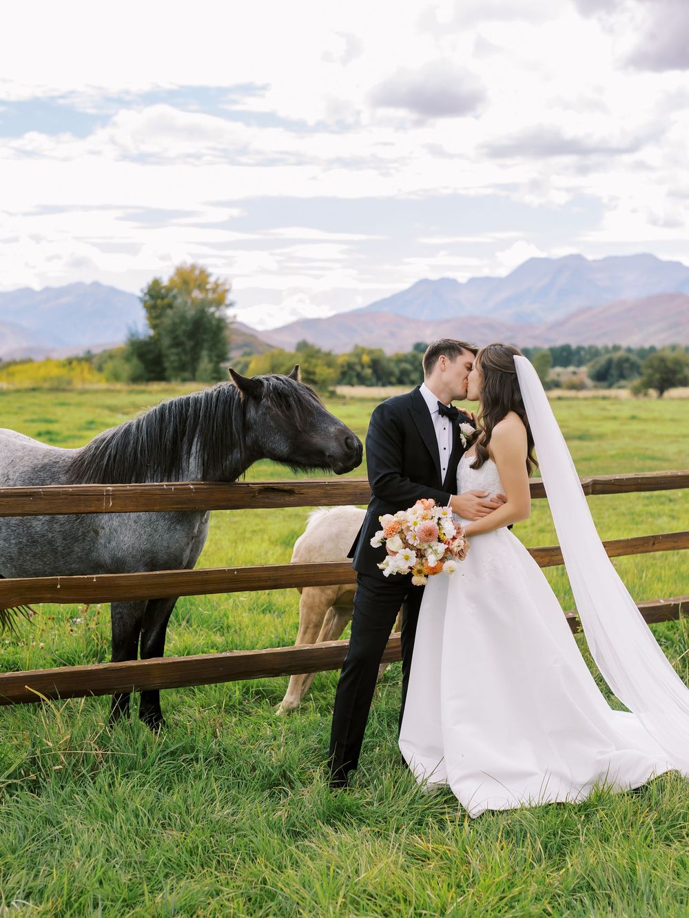 Bride and groom share a romantic moment by a wooden fence with horses and mountain backdrop on their wedding day.