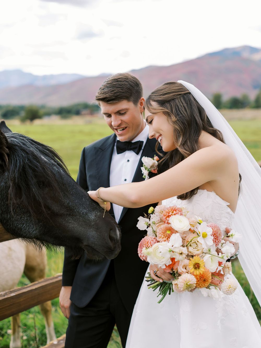 Smiling bride and groom pet a black horse while bride holds colorful bouquet at outdoor wedding.