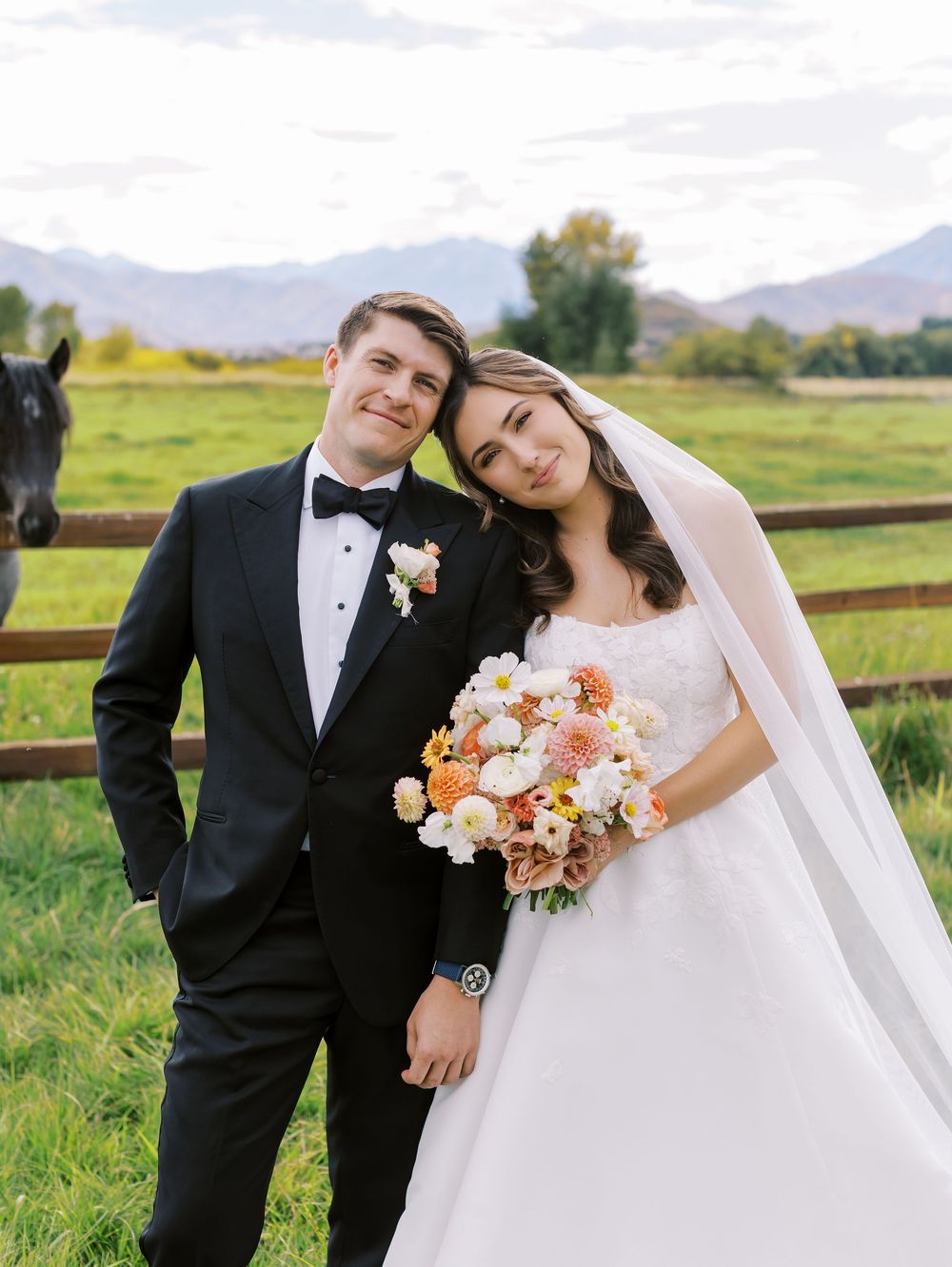 Bride and groom pose together in a field with mountain backdrop, she holds a peach bouquet, he wears a black tuxedo.