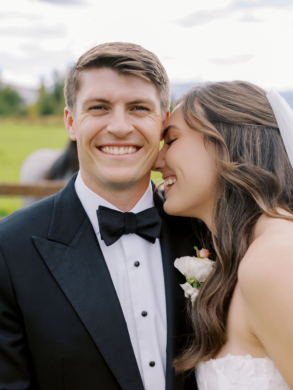 Smiling groom in black tuxedo and bow tie with bride in white wedding dress sharing a joyful moment outdoors.