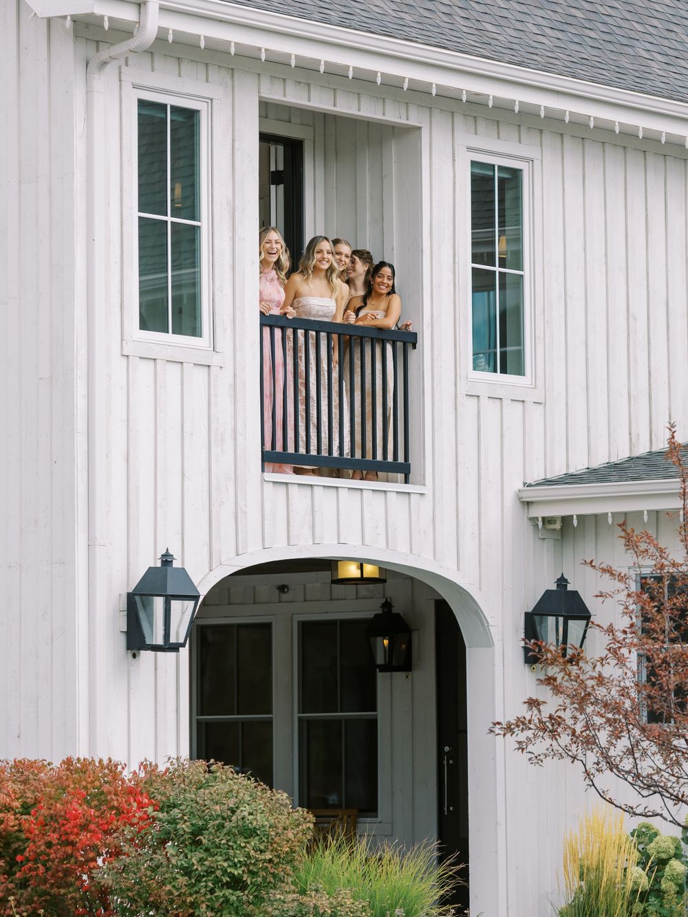 Group of women smiling from a white farmhouse balcony above an arched entryway with black lantern lights.