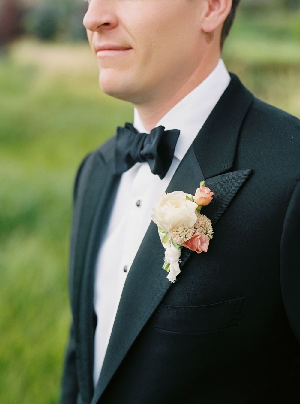 Groom in black tuxedo with bow tie wearing pink and white floral boutonniere on lapel.