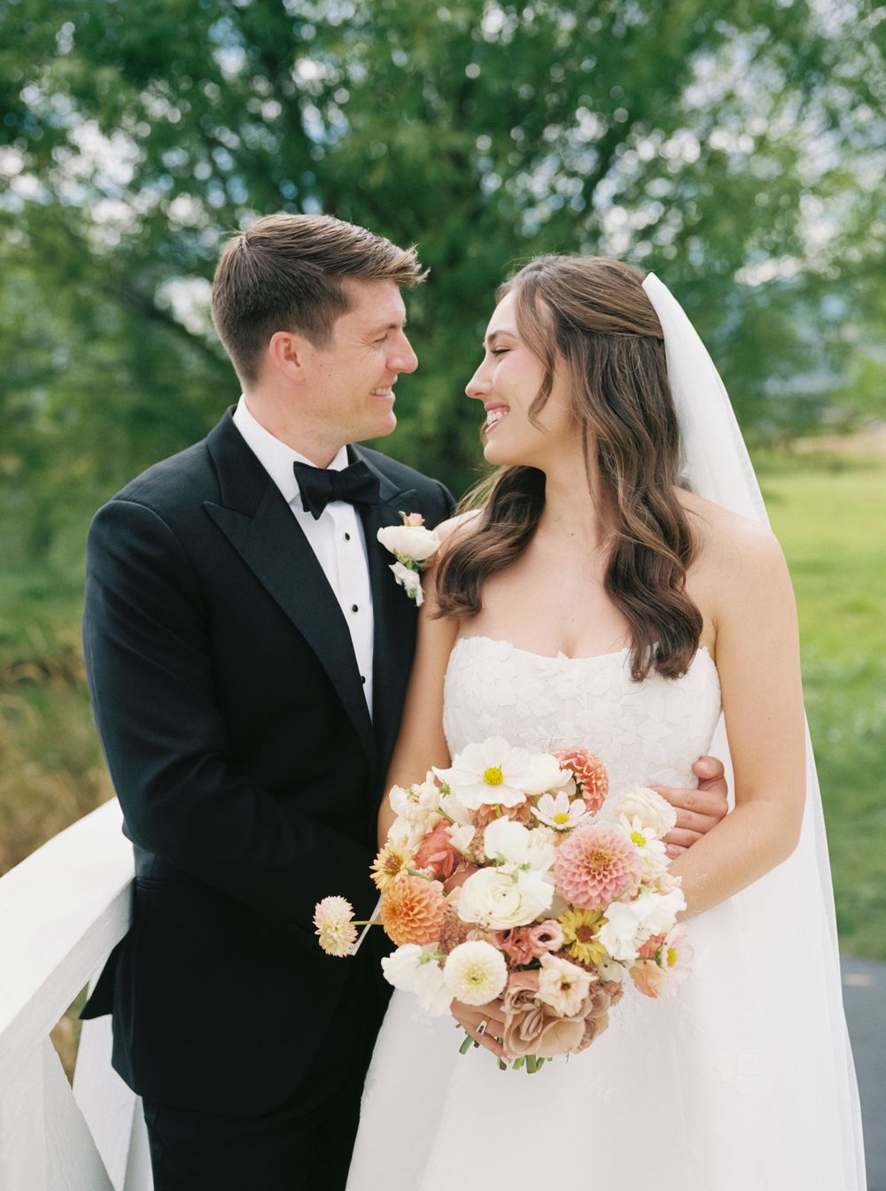 Bride and groom smiling at each other outdoors, she holds a peach and white bouquet, he wears a black tuxedo.