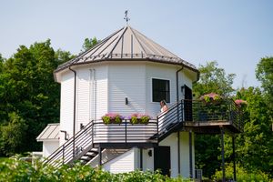 White octagonal tower building with metal roof, wraparound deck with flower boxes, surrounded by lush green trees on a sunny day.