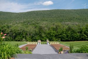 Outdoor wedding ceremony setup with wooden chairs on a green lawn, framed by lush forested mountains under a blue sky.