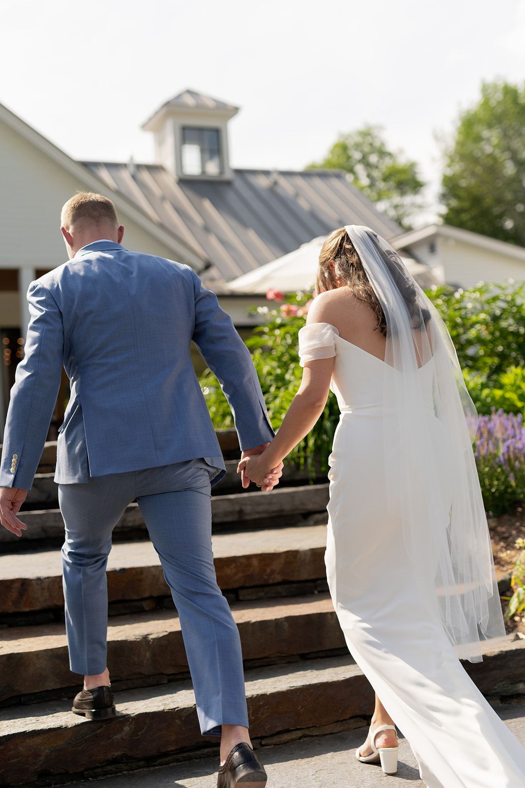 Bride and groom holding hands walking up stone steps toward a white barn venue on a sunny wedding day.