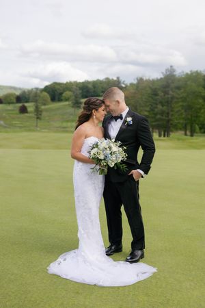 Bride and groom share a romantic moment on a golf course, bride holding a white and blue bouquet in a lace gown.