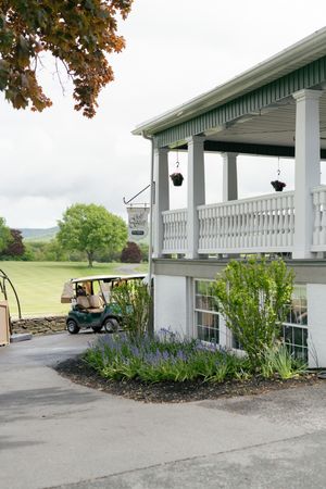 A golf cart parked beside a charming two-story clubhouse with white balcony railings, surrounded by lush greenery and lavender plants.
