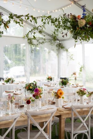 Elegant wedding reception tent with lush greenery ceiling decor, string lights, white chairs, and colorful floral centerpieces.