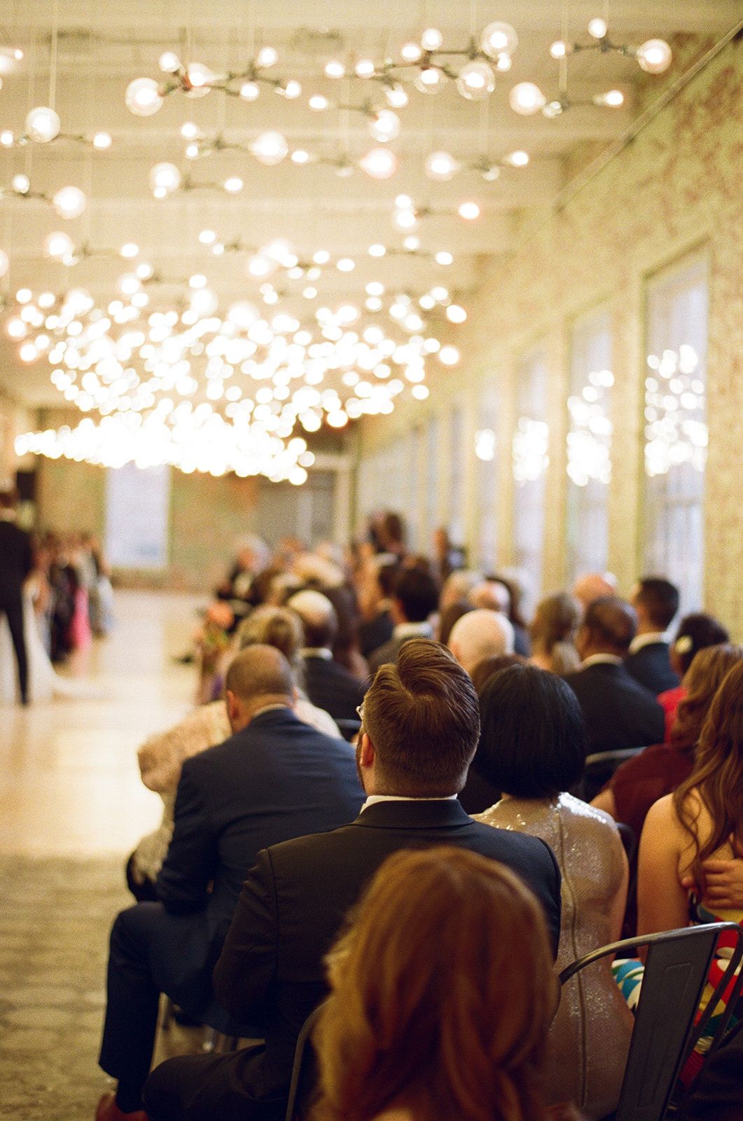 Guests seated in elegant venue with globe string lights overhead during a wedding ceremony.
