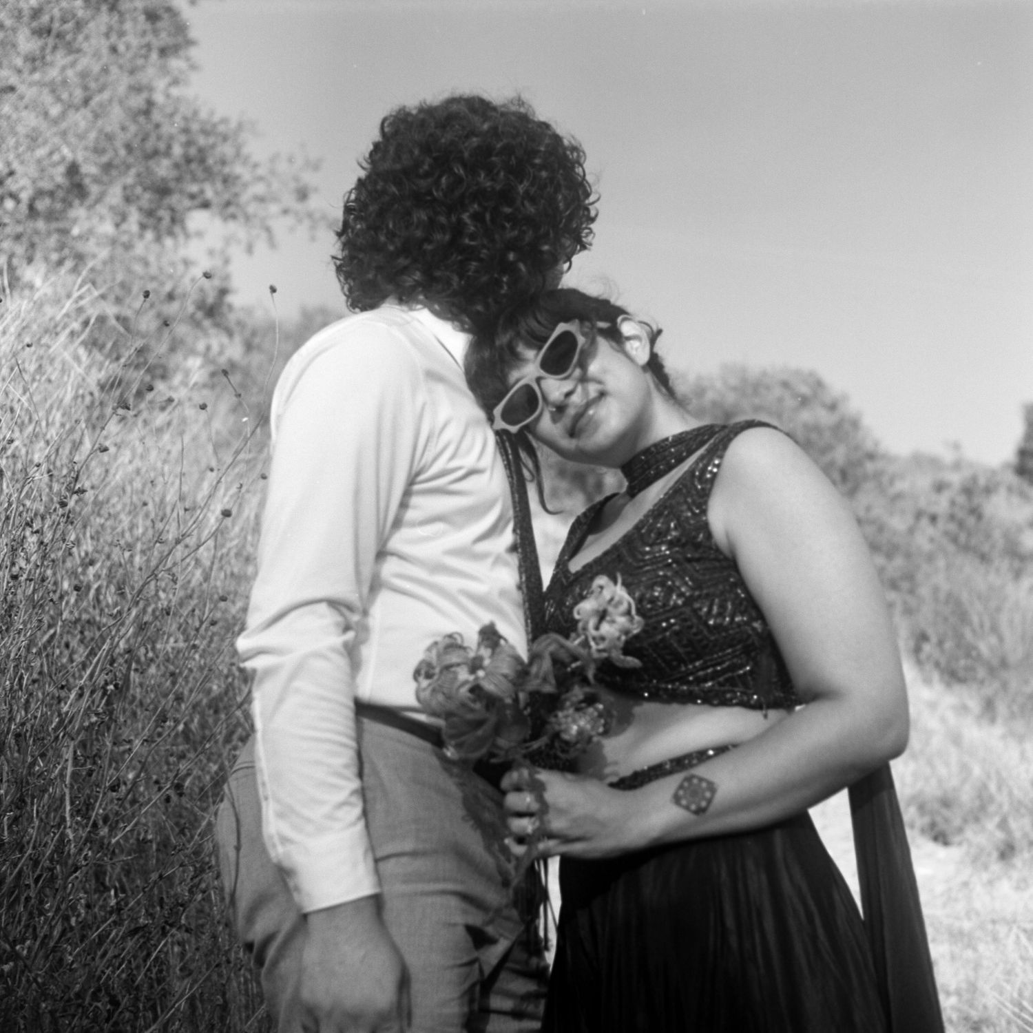 Couple embracing outdoors in black and white photo, woman in sequin dress holding flowers, man in white shirt.