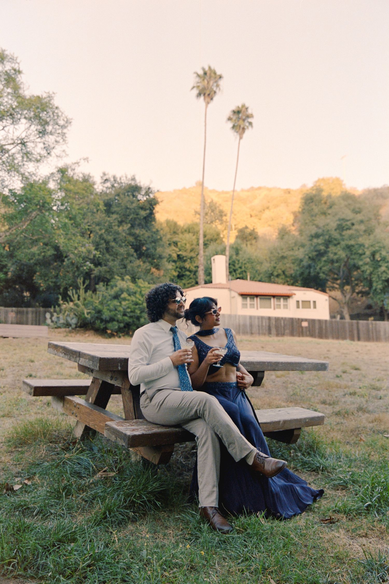Couple sitting together at a wooden picnic table outdoors, with palm trees and golden hills in the background.