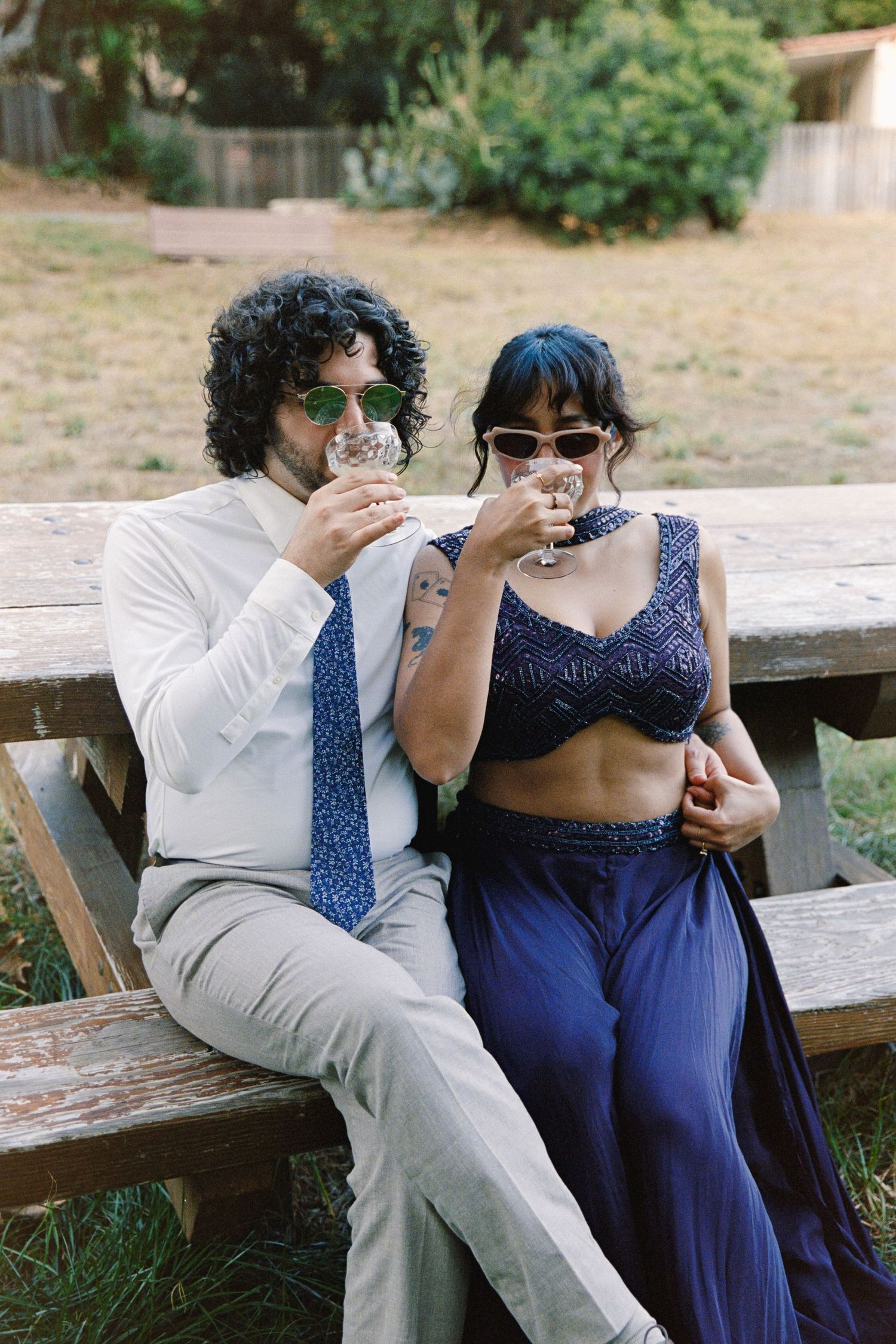 Couple in formal attire sipping wine on a wooden bench outdoors at a garden event.