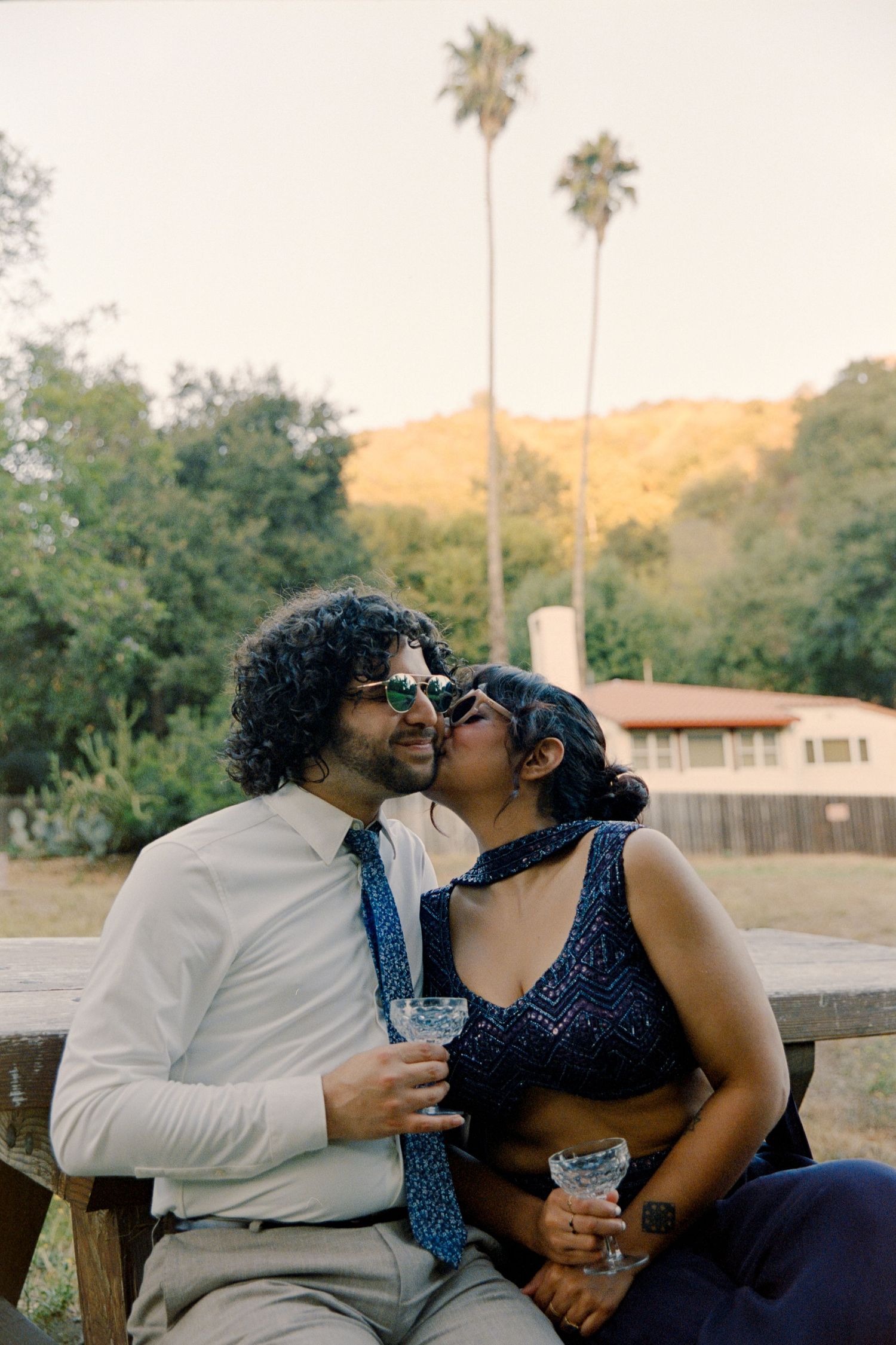Couple sharing a kiss while holding cocktails outdoors, with palm trees and hills in the background.