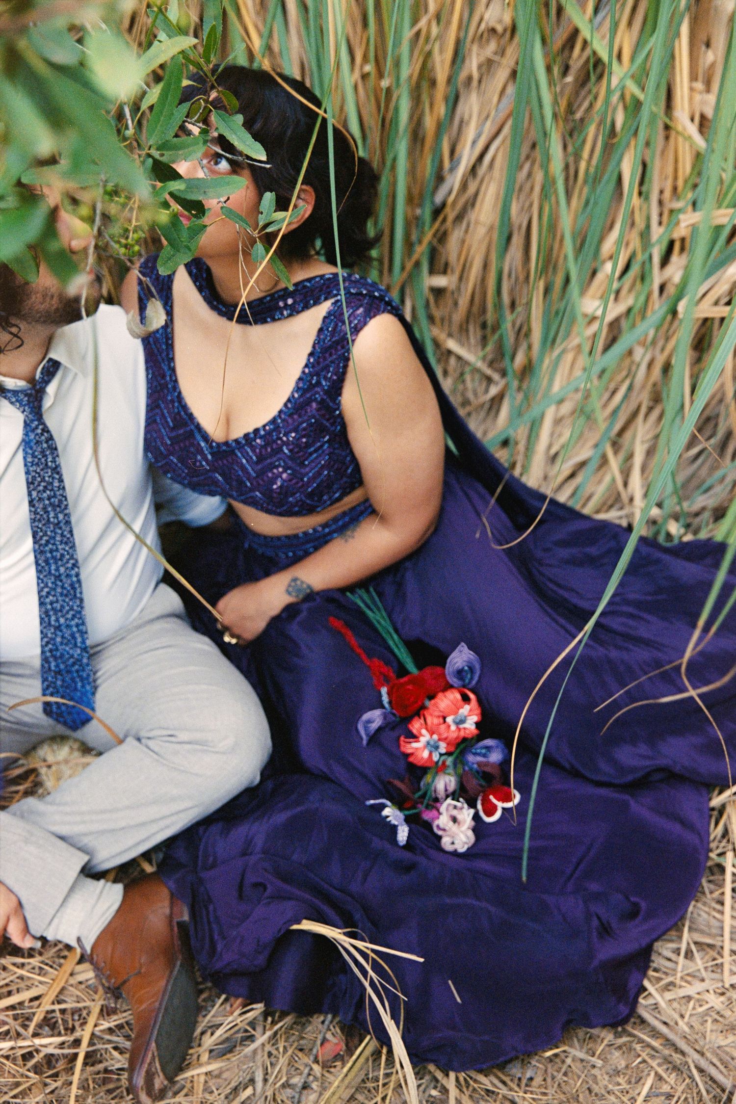Couple sitting outdoors among tall grass, woman in navy blue embroidered lehenga holding colorful bouquet, man in white shirt and blue tie.