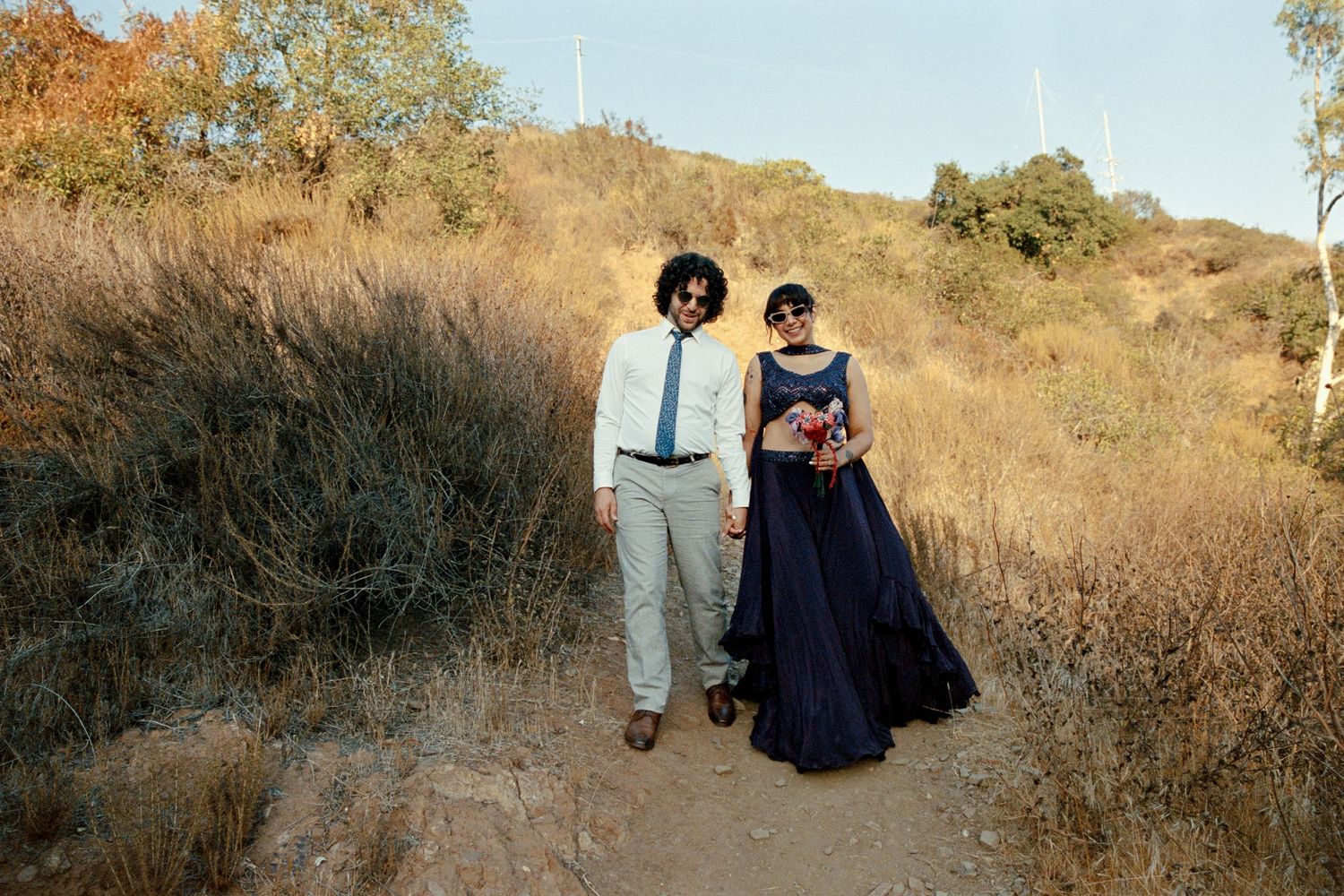 Couple in formal attire walking on a dirt trail surrounded by dry brush and hills on a sunny day.