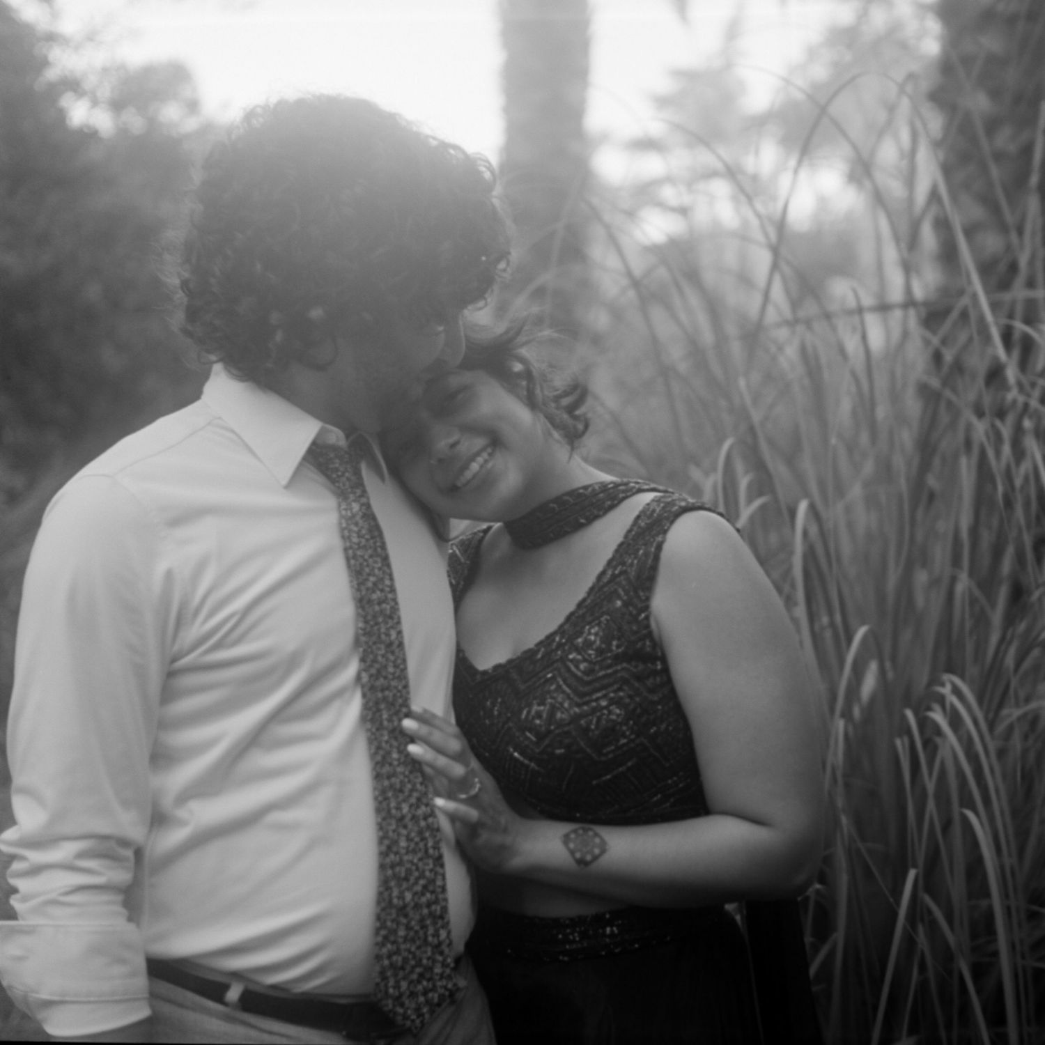 Black and white romantic photo of couple embracing near tall grass, man in white shirt and tie kissing womans cheek.