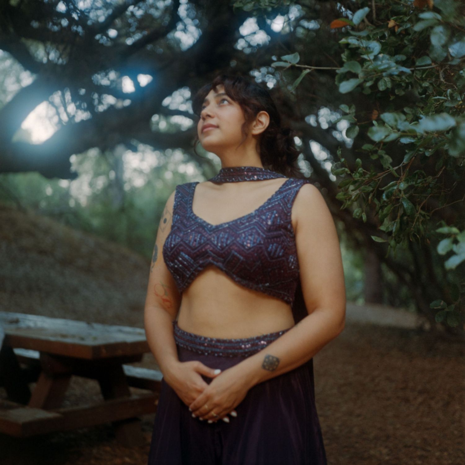 Woman in ornate purple beaded crop top and skirt standing under a large tree outdoors.