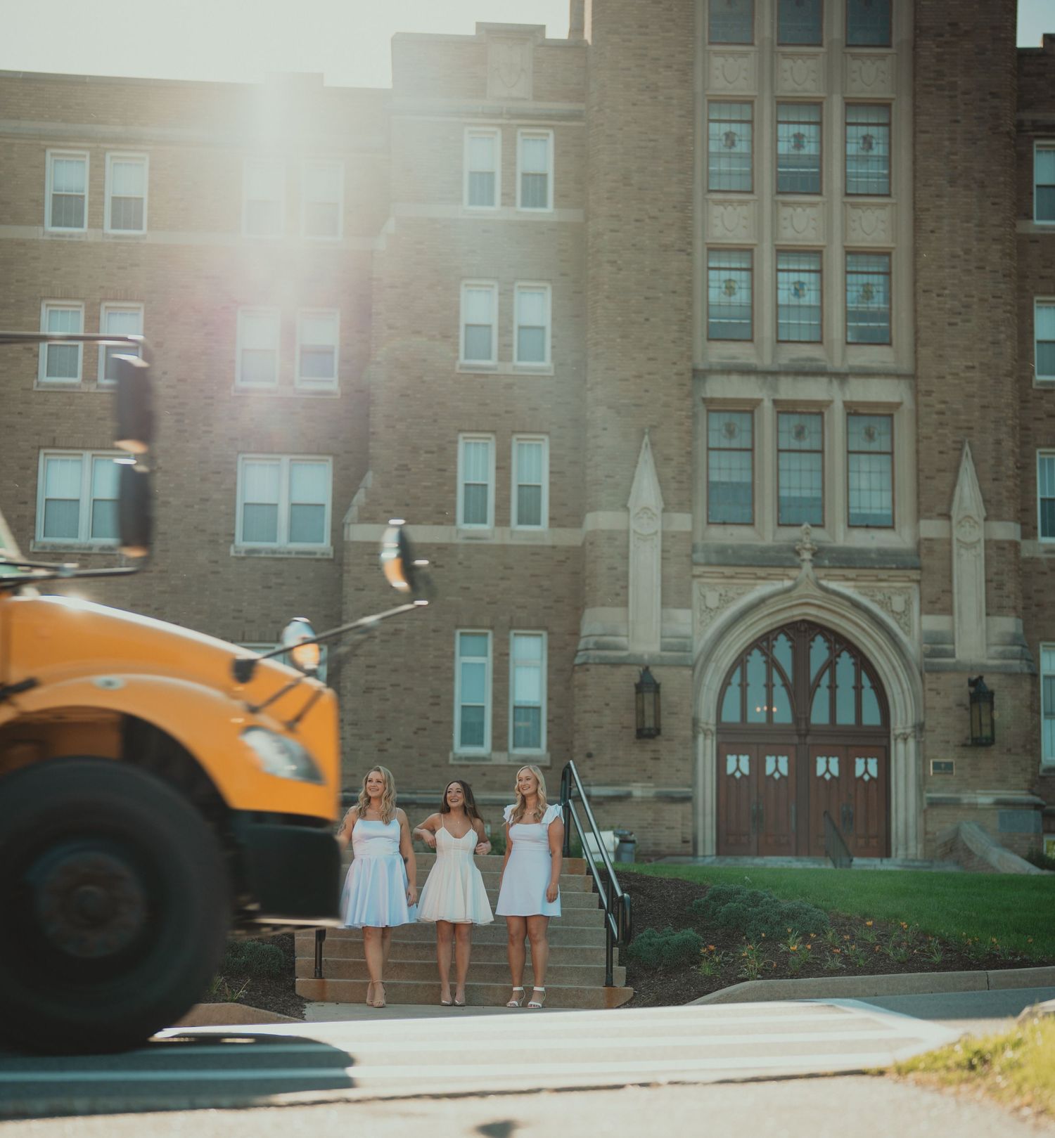 Three women in white dresses stand before a large brick building as a yellow school bus passes by in golden sunlight.