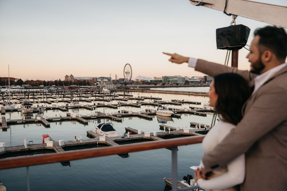 Couple on ship deck overlooking Chicago Navy Pier marina at sunset, man pointing at waterfront with Ferris wheel in background.