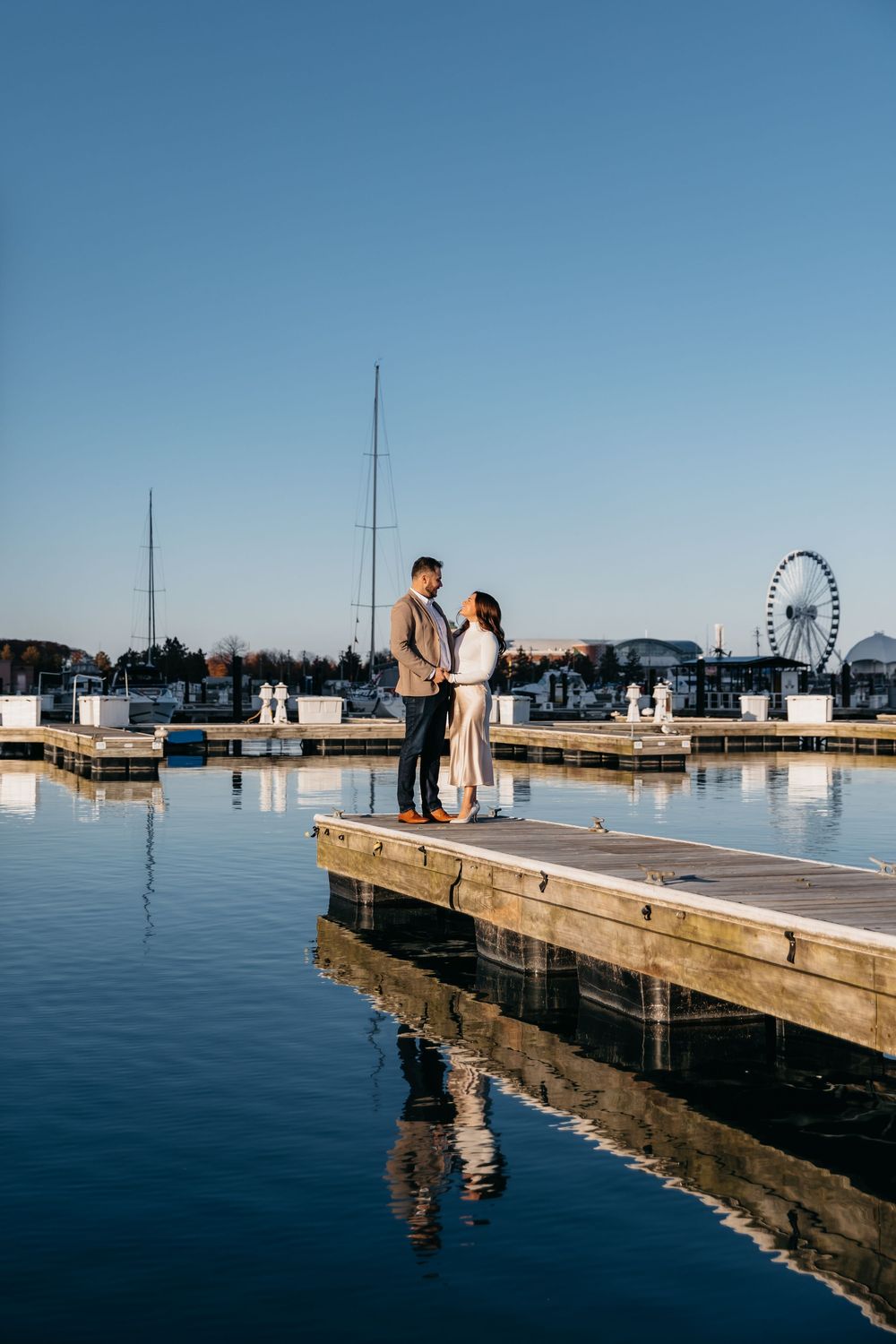 Couple standing during engagement on a wooden dock at Chicago Navy Pier, with calm water reflections and a Ferris wheel in the background.