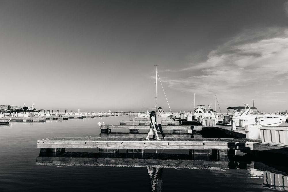 Black and white photo of Couple walking during engagement session at Chicago Navy Pier, with calm water reflections and boats on background