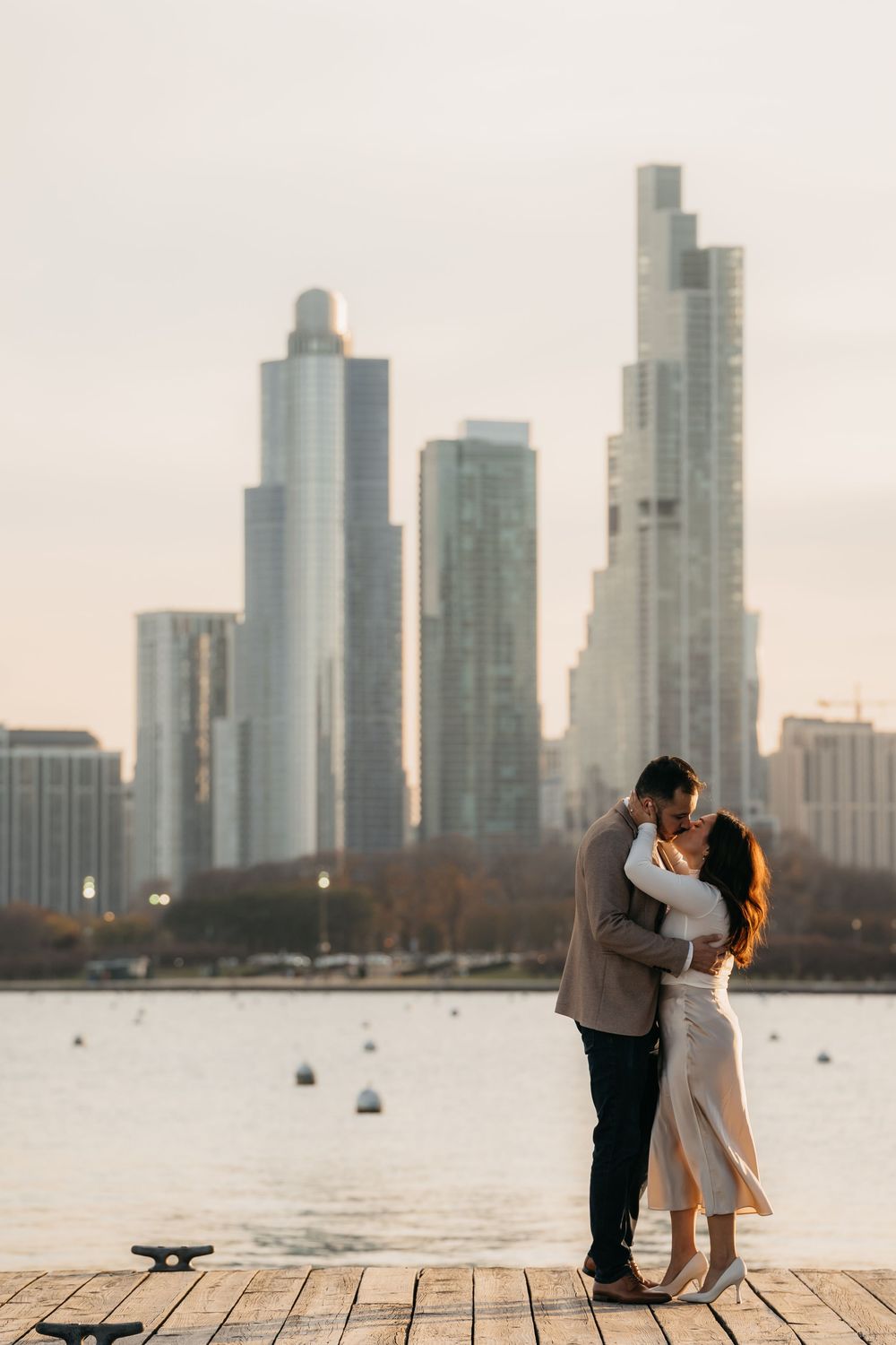 Couple sharing a romantic kiss on a waterfront Navy pier with Chicago city skyline skyscrapers in the background at sunset.