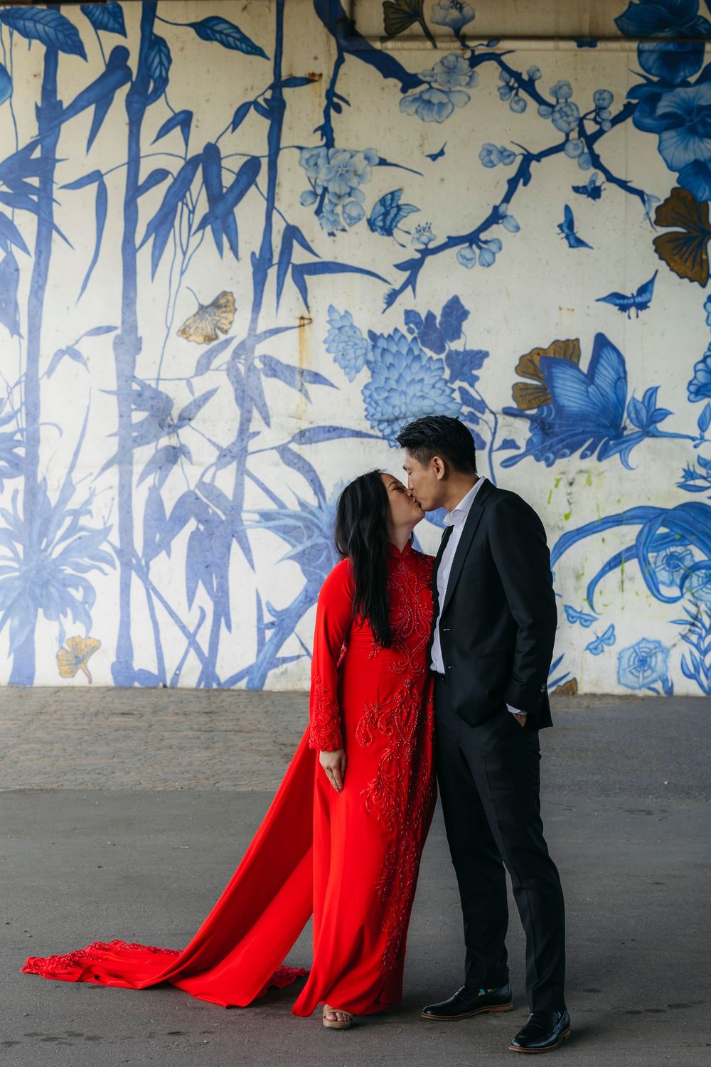 Couple kissing in red ao dai and black suit before blue floral mural wall in Ping Tom Park in Chinatown Chicago during engagement session