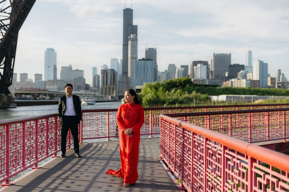 Couple in red traditional attire poses on red bridge in Ping Tom Park in Chinatown with Chicago skyline in the background during engagement