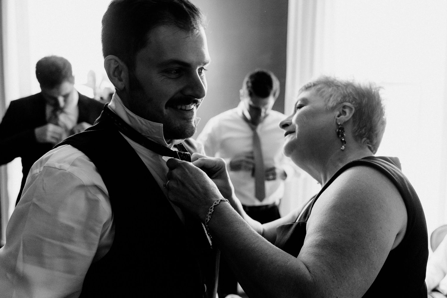 Filmic wedding photo of Milwaukee groom's mother fixing his tie as groomsmen get ready in the background.