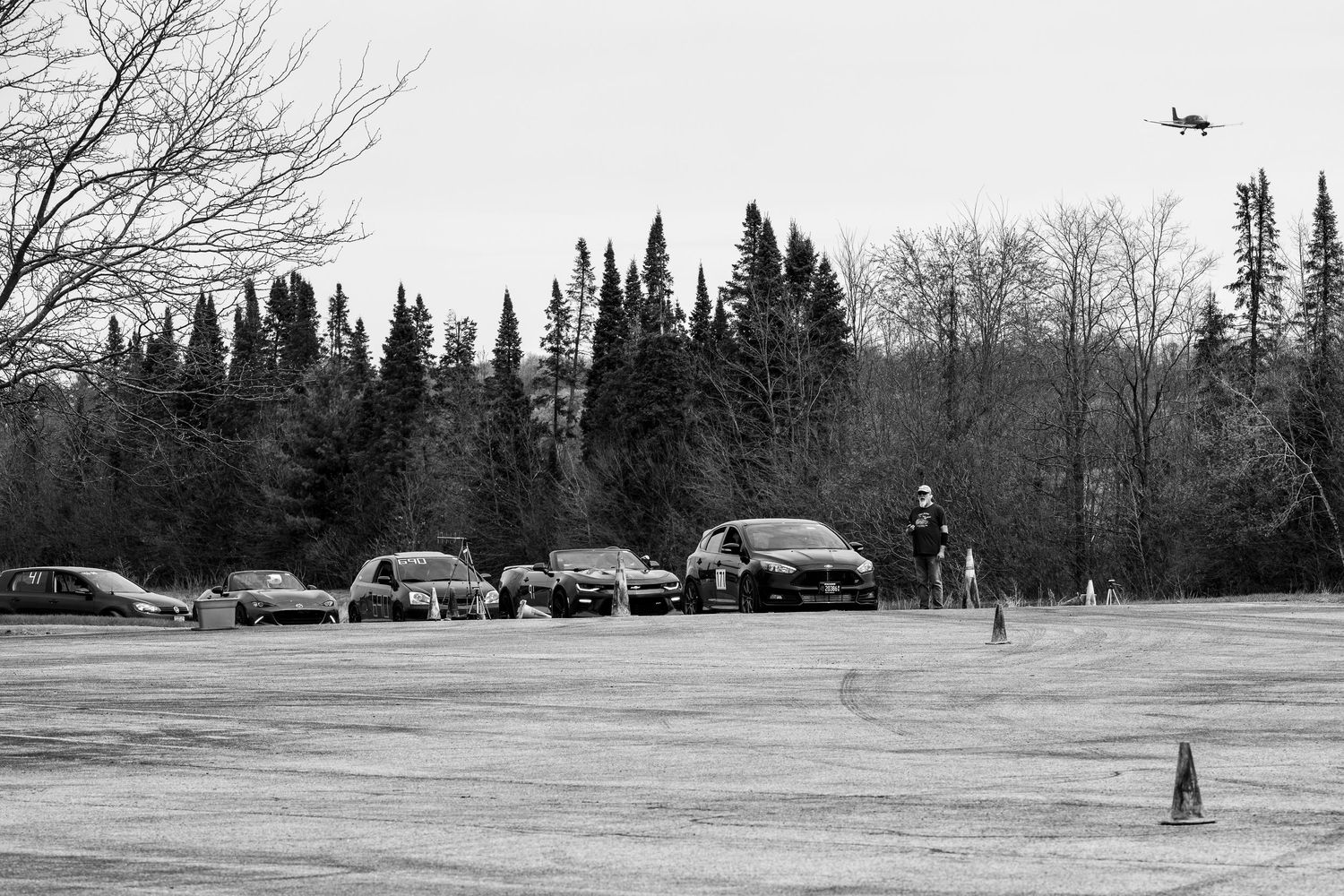 Black and white photo of cars parked in a lot surrounded by tall evergreen trees during an outdoor driving event.