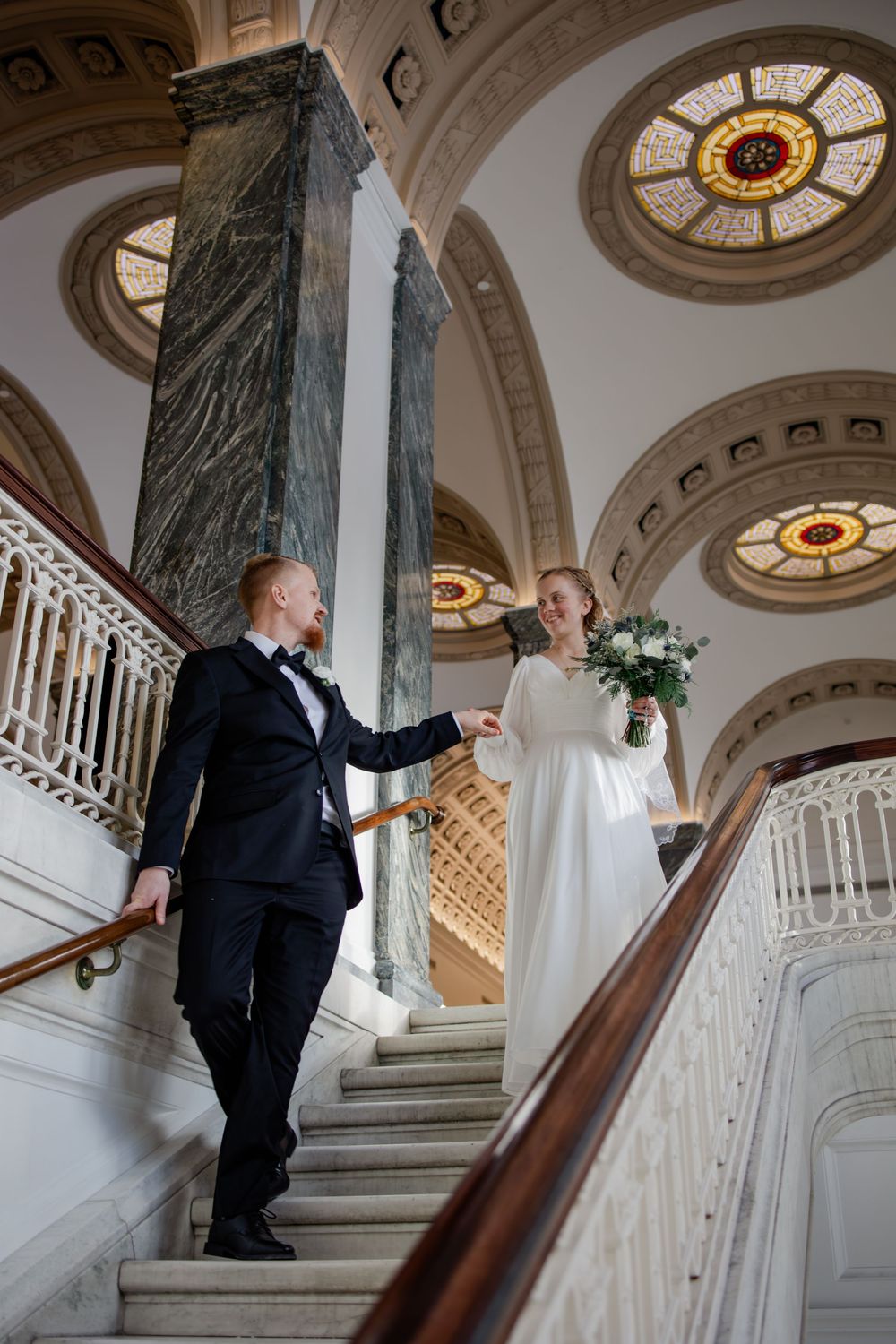 Bride and groom holding hands on grand marble staircase in elegant venue with stained glass ceiling.