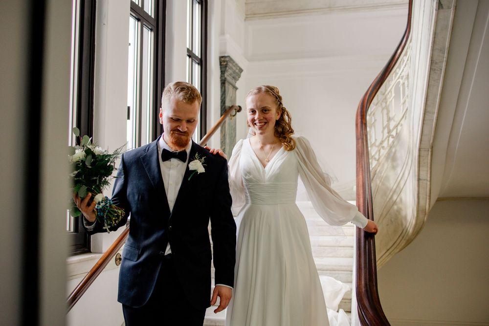 Bride and groom walking down elegant staircase, smiling, bride in white gown, groom in navy suit with bow tie.