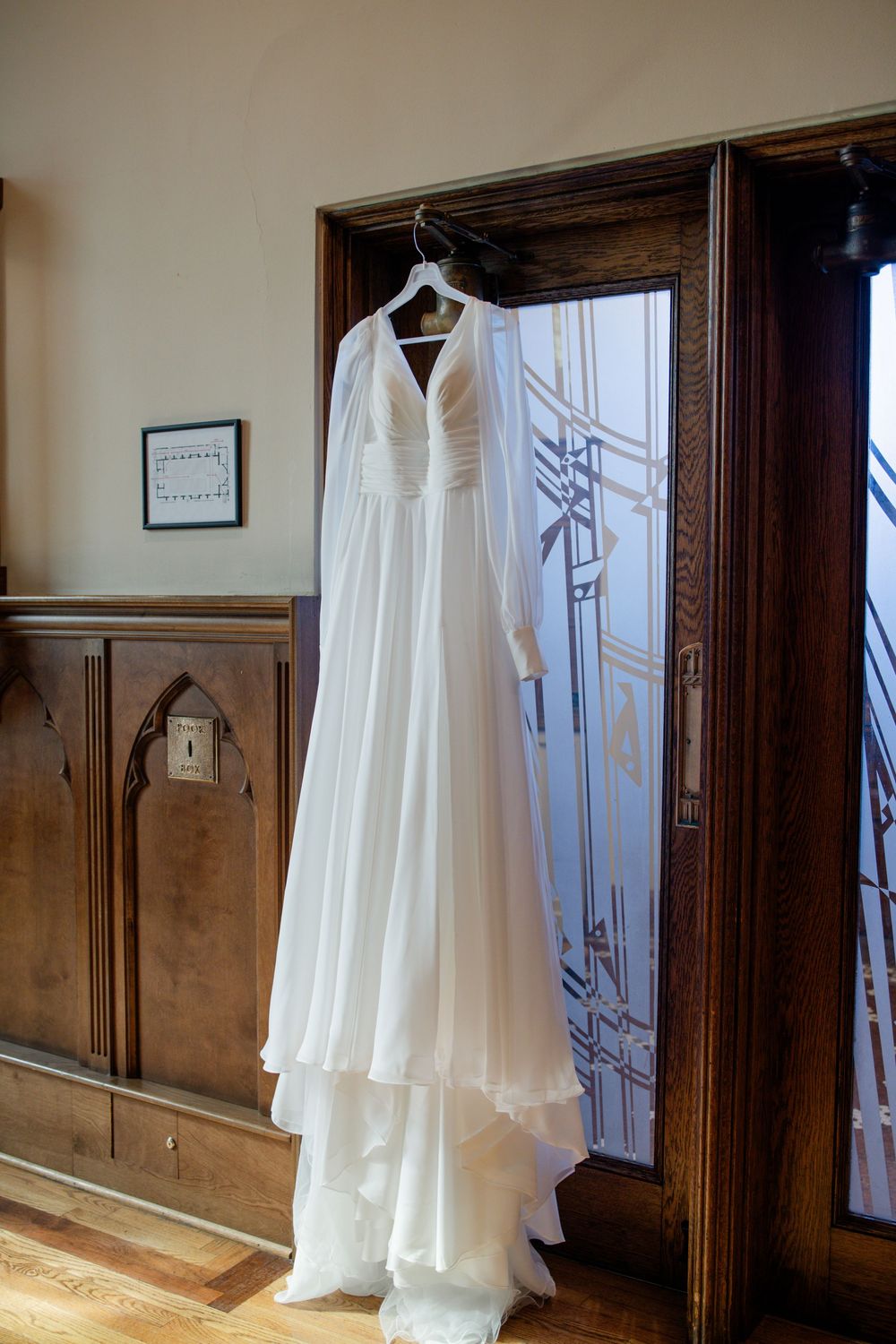 Elegant white bridal gown with flowing chiffon layers hanging on a wooden door in a classic venue.