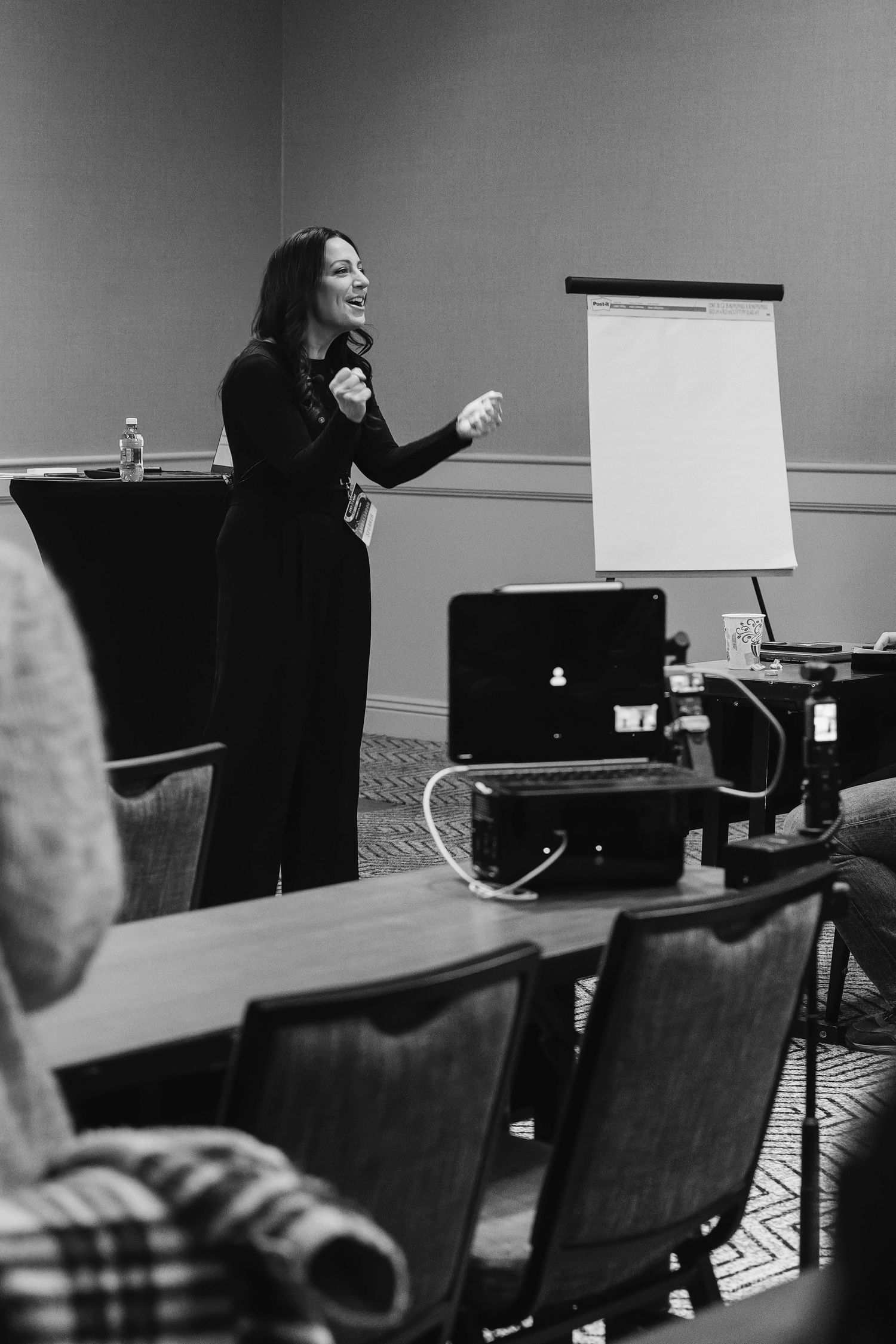 Woman in black outfit presenting at a conference, gesturing near a flip chart with laptops on tables.