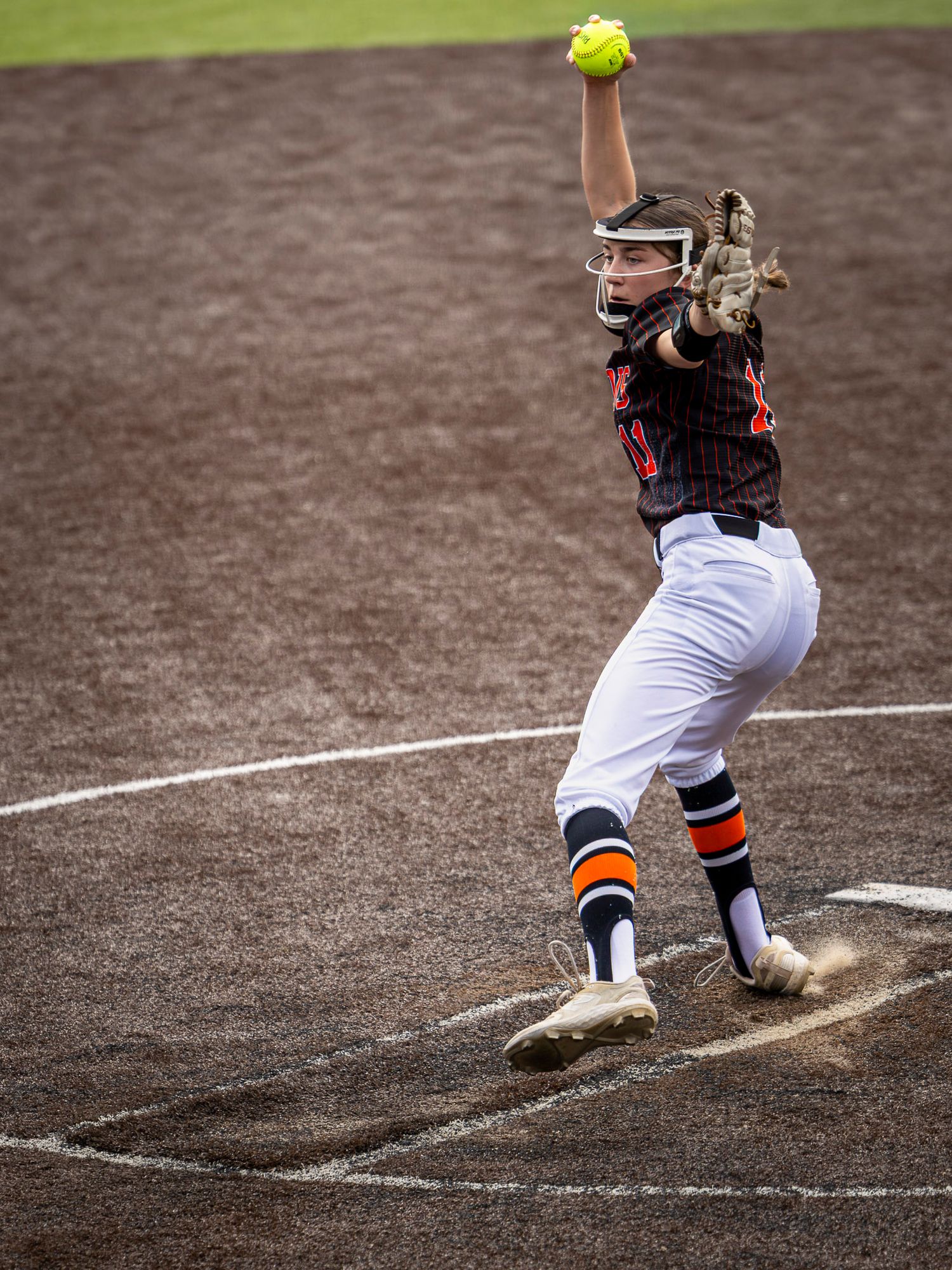 Gravette softball pitcher in orange and black uniform winds up to throw a yellow ball during a game.