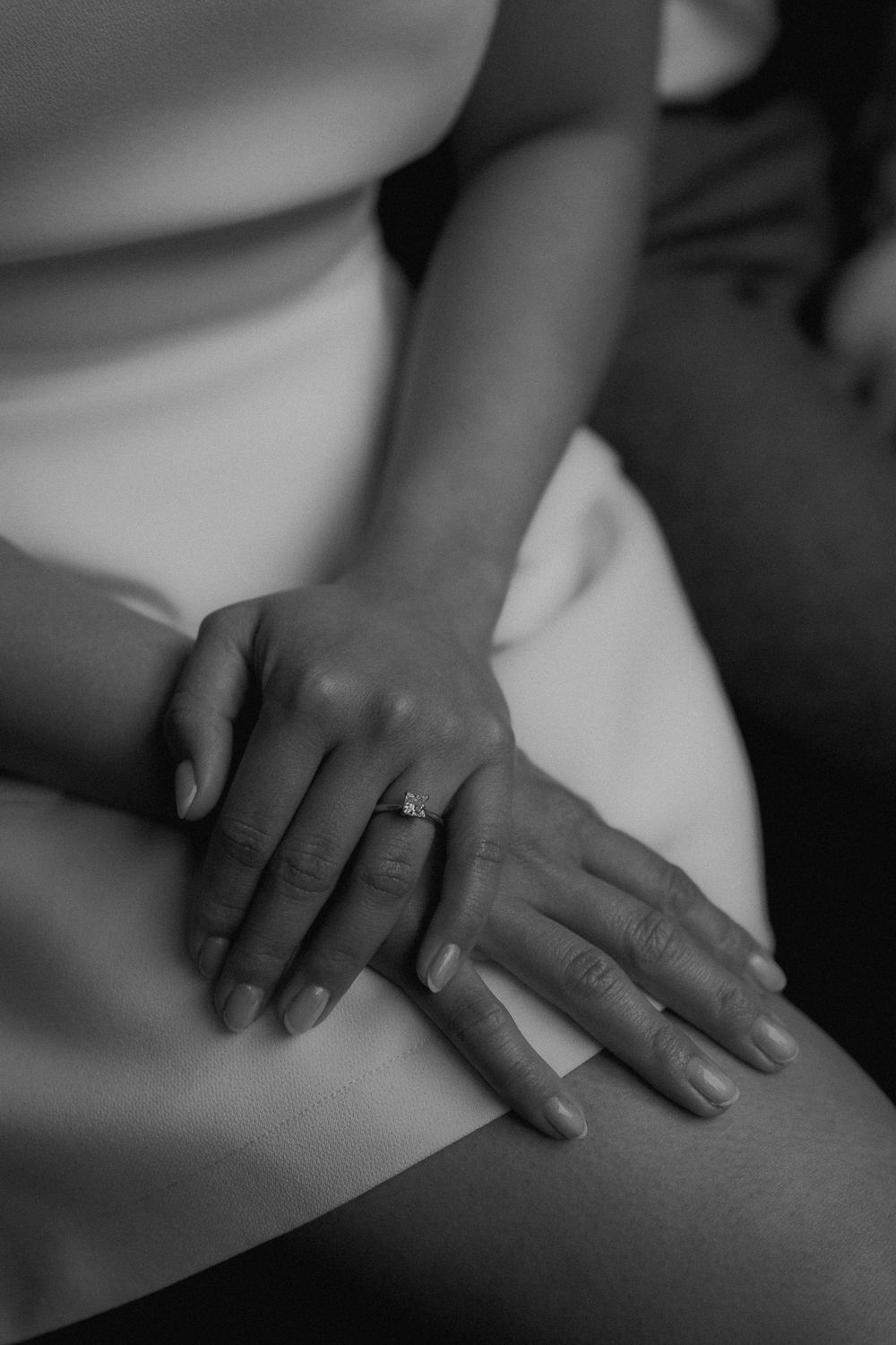 Close-up black and white photo of a bride's hands resting on her lap, showcasing her elegant diamond engagement ring.