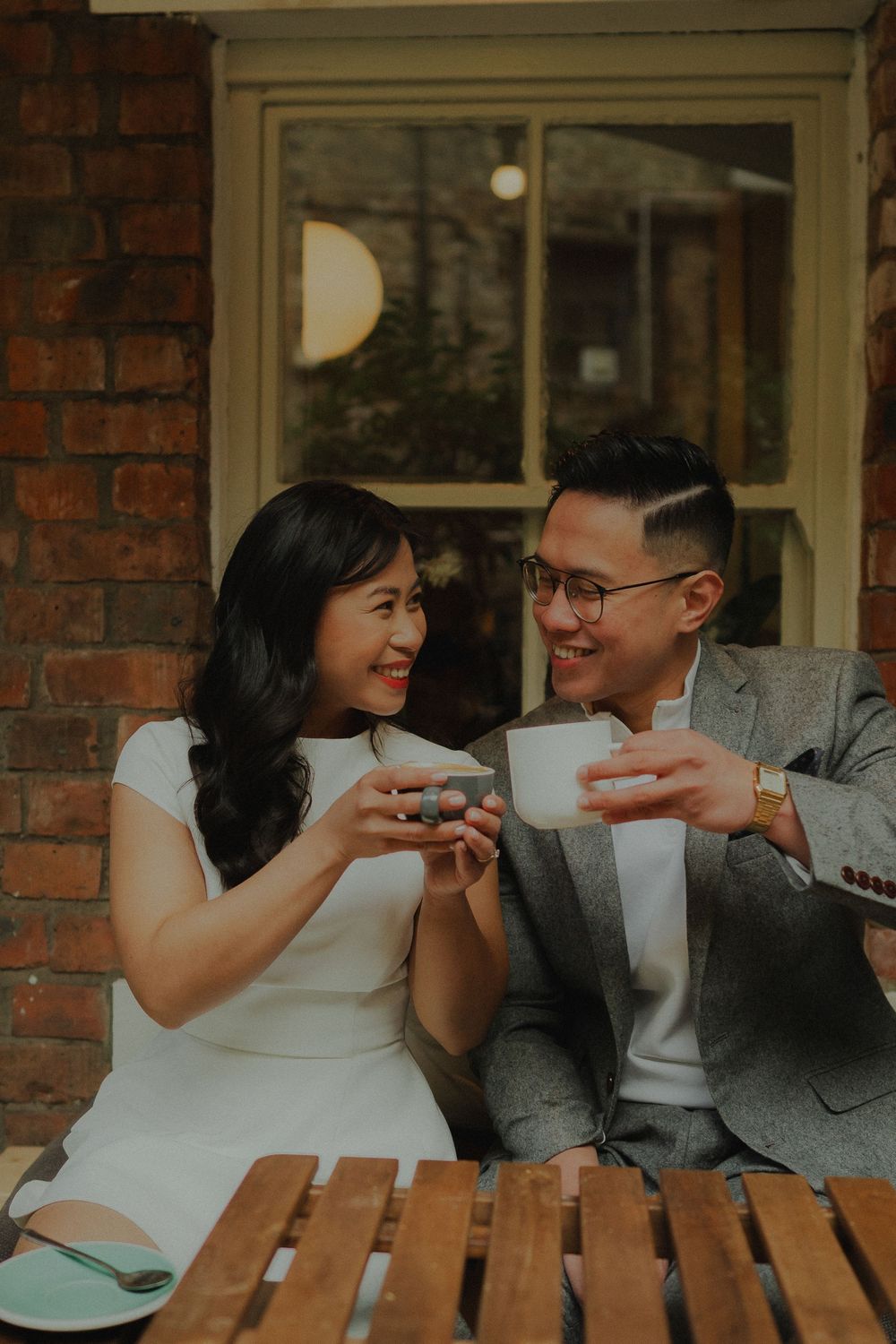 A smiling Asian couple toasting with coffee cups at an outdoor cafe table, dressed in elegant white and grey attire.