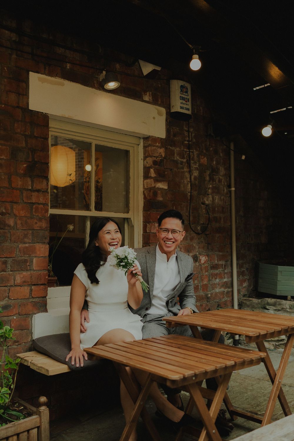 A smiling couple in wedding attire sits at a wooden table outside a brick building, the bride holding a small bouquet.