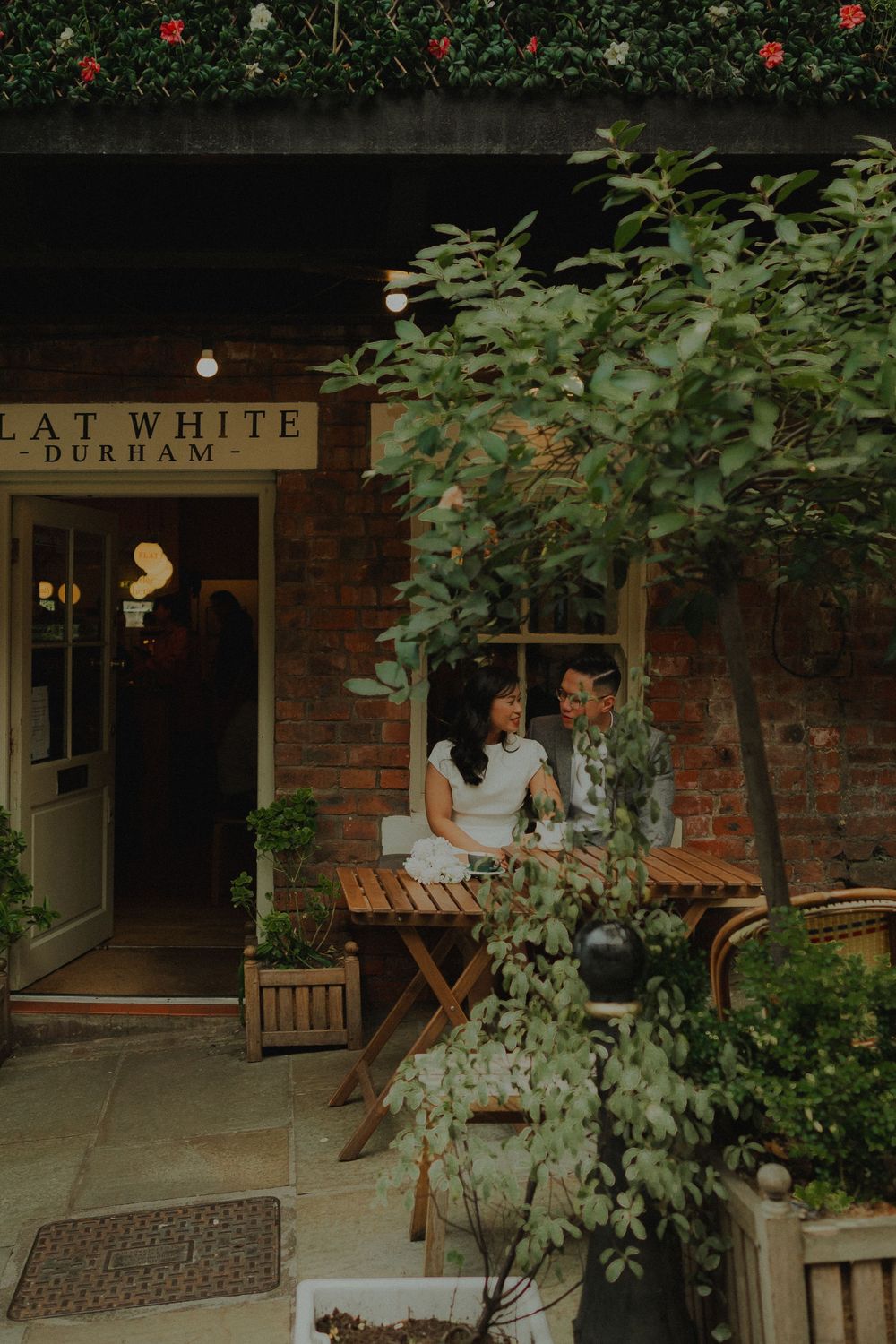 Couple sitting at outdoor wooden table at Flat White Durham cafe, surrounded by lush greenery and brick walls.