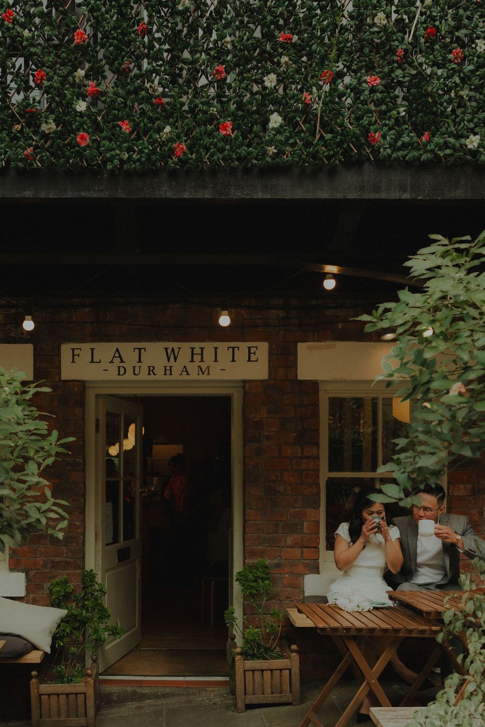 Couple enjoying coffee outside Flat White Durham cafe with brick walls, fairy lights, and lush green ivy facade.
