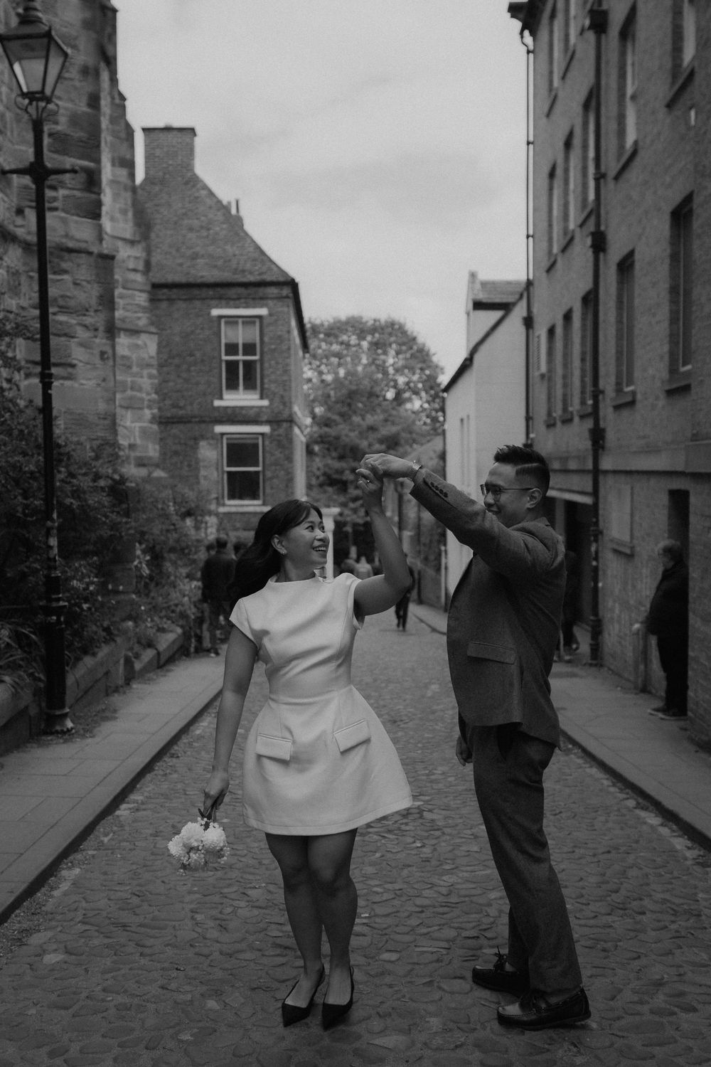 Black and white photo of a couple dancing on a cobblestone street, bride in short dress holding bouquet, groom in suit.